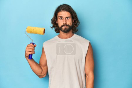 Photo for Bearded man painting with a yellow roller confused, feels doubtful and unsure. - Royalty Free Image