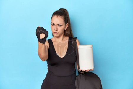 Foto de Mujer joven sosteniendo la botella de proteína en un entorno deportivo que muestra el puño a la cámara, expresión facial agresiva. - Imagen libre de derechos
