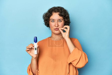 Photo for Young woman holding pregnancy test, studio background with fingers on lips keeping a secret. - Royalty Free Image