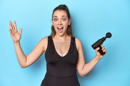 Photo for Active woman with an electric massager in a blue studio receiving a pleasant surprise, excited and raising hands. - Royalty Free Image