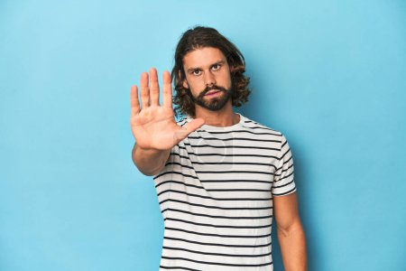 Téléchargez les photos : Homme barbu dans une chemise rayée, fond bleu debout avec la main tendue montrant le signe d'arrêt, vous empêchant. - en image libre de droit