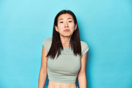 Photo for Asian woman in summer green top, studio backdrop, shrugs shoulders and open eyes confused. - Royalty Free Image