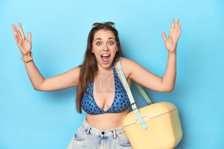 Photo for Woman in bikini with a portable beach cooler, blue studio receiving a pleasant surprise, excited and raising hands. - Royalty Free Image