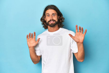 Téléchargez les photos : Homme barbu dans une chemise blanche, fond bleu rejetant quelqu'un montrant un geste de dégoût. - en image libre de droit