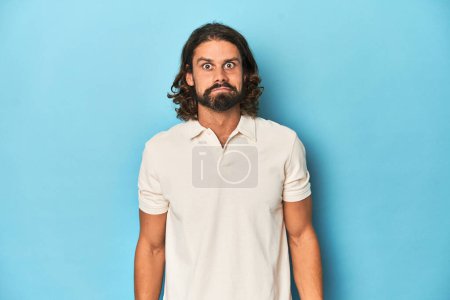 Téléchargez les photos : Homme aux cheveux longs dans un polo blanc, studio bleu haussant les épaules et les yeux ouverts confus. - en image libre de droit