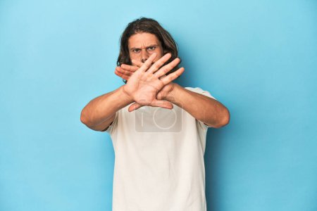 Téléchargez les photos : Homme aux cheveux longs dans un polo blanc, studio bleu faisant un geste de déni - en image libre de droit