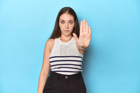 Photo for Woman in a striped top posing on a blue studio backdrop standing with outstretched hand showing stop sign, preventing you. - Royalty Free Image