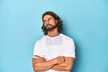 Photo for Bearded man in a white shirt, blue backdrop tired of a repetitive task. - Royalty Free Image