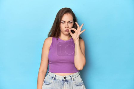 Téléchargez les photos : Jeune femme à la mode dans un haut violet sur fond bleu avec les doigts sur les lèvres gardant un secret. - en image libre de droit