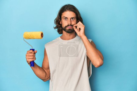Photo for Bearded man painting with a yellow roller with fingers on lips keeping a secret. - Royalty Free Image