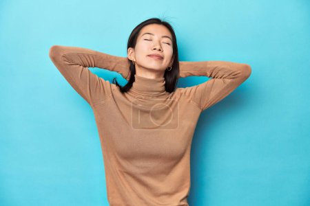 Photo for Young Asian woman in brown turtleneck, feeling confident, with hands behind the head. - Royalty Free Image