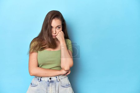 Téléchargez les photos : Jeune femme caucasienne dans un haut vert sur un fond bleu qui se sent triste et coûteux, en regardant l'espace de copie. - en image libre de droit
