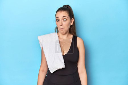Photo for Fit young woman with a towel on her shoulder, studio shot shrugs shoulders and open eyes confused. - Royalty Free Image