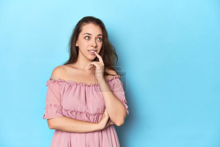 Téléchargez les photos : Jeune femme portant une robe rose sur fond de studio bleu regardant latéralement avec une expression douteuse et sceptique. - en image libre de droit