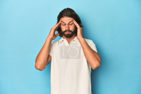 Foto de Hombre de pelo largo en un polo blanco, estudio azul tocando templos y teniendo dolor de cabeza. - Imagen libre de derechos