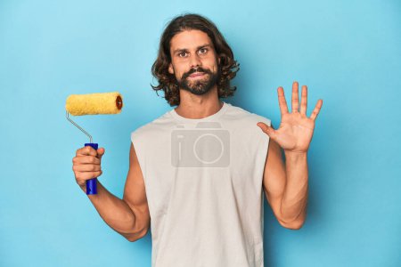 Photo for Bearded man painting with a yellow roller smiling cheerful showing number five with fingers. - Royalty Free Image