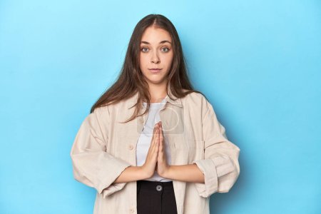 Foto de Mujer joven con estilo en una camiseta sobre un fondo azul rezando, mostrando devoción, persona religiosa en busca de inspiración divina. - Imagen libre de derechos