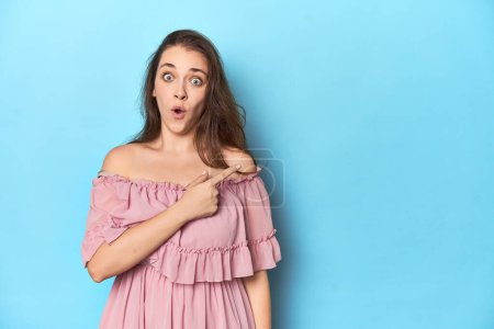 Téléchargez les photos : Jeune femme portant une robe rose sur fond de studio bleu pointant vers le côté - en image libre de droit