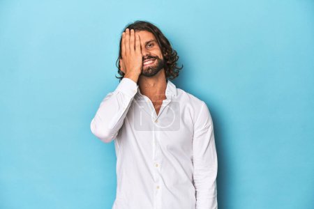Photo for Bearded man in a white shirt, blue backdrop having fun covering half of face with palm. - Royalty Free Image
