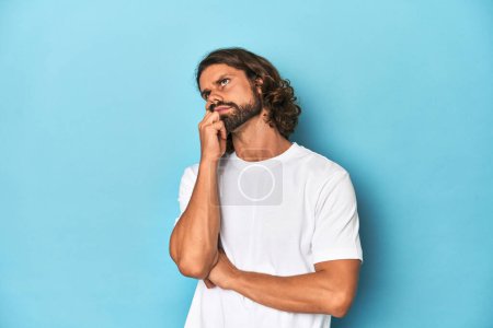 Photo for Bearded man in a white shirt, blue backdrop thinking and looking up, being reflective, contemplating, having a fantasy. - Royalty Free Image
