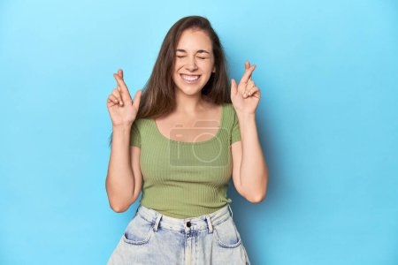 Téléchargez les photos : Jeune femme caucasienne dans un haut vert sur un fond bleu croisant les doigts pour avoir de la chance - en image libre de droit