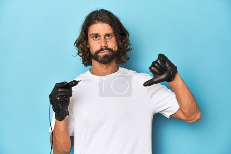 Photo for Bearded tattooist with tattoo machine at work showing a dislike gesture, thumbs down. Disagreement concept. - Royalty Free Image