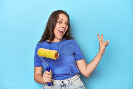 Téléchargez les photos : Jeune femme avec rouleau de peinture jaune sur un fond bleu joyeux et insouciant montrant un symbole de paix avec les doigts. - en image libre de droit