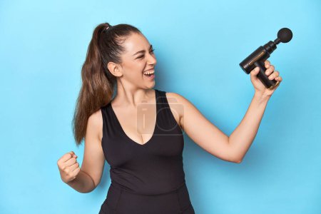 Foto de Mujer activa con un masajeador eléctrico en un estudio azul levantando puño después de una victoria, concepto ganador. - Imagen libre de derechos