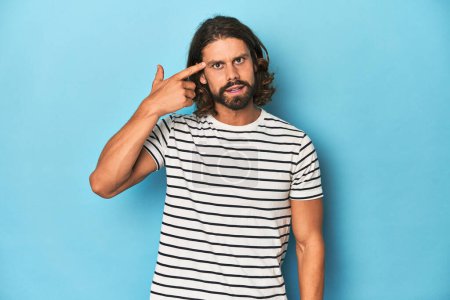 Photo for Bearded man in a striped shirt, blue backdrop showing a disappointment gesture with forefinger. - Royalty Free Image
