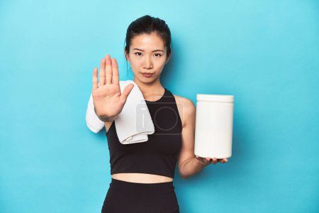 Photo for Young Asian sportswoman with protein bottle, gym setup, standing with outstretched hand showing stop sign, preventing you. - Royalty Free Image