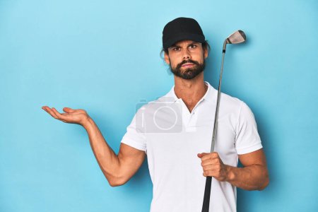 Foto de Golfista de pelo largo con palo y sombrero mostrando un espacio de copia en una palma y sosteniendo otra mano en la cintura. - Imagen libre de derechos
