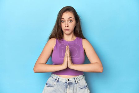 Foto de Mujer joven de moda en una parte superior púrpura sobre fondo azul rezando, mostrando devoción, persona religiosa en busca de inspiración divina. - Imagen libre de derechos