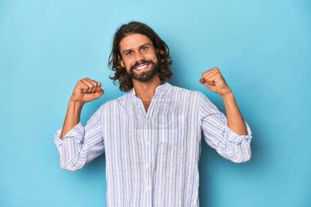 Téléchargez les photos : Homme barbe à rayures bleues chemise, studio bleu célébrant une victoire, passion et enthousiasme, expression heureuse. - en image libre de droit