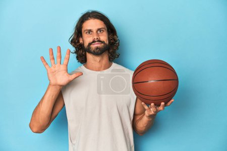 Téléchargez les photos : Homme barbu avec basket-ball en studio bleu souriant gai montrant numéro cinq avec les doigts. - en image libre de droit