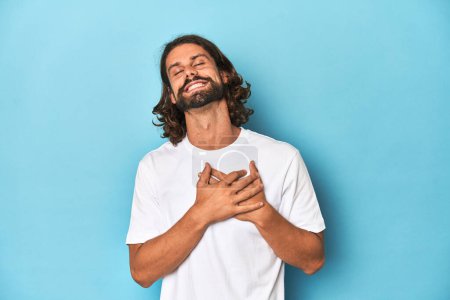 Foto de Hombre barbudo en una camisa blanca, fondo azul riendo manteniendo las manos en el corazón, concepto de felicidad. - Imagen libre de derechos