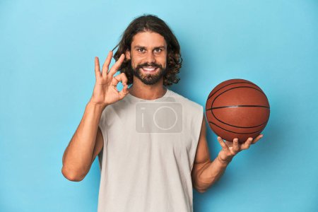 Téléchargez les photos : Homme barbu avec basket-ball en studio bleu joyeux et confiant montrant geste ok. - en image libre de droit