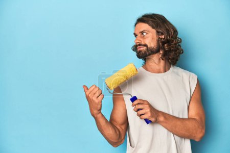 Photo for Bearded man painting with a yellow roller points with thumb finger away, laughing and carefree. - Royalty Free Image