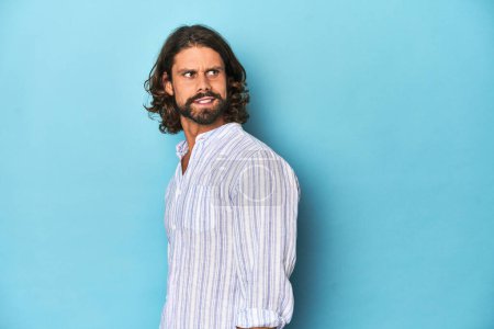 Photo for Man with beard in blue striped shirt, blue studio looks aside smiling, cheerful and pleasant. - Royalty Free Image