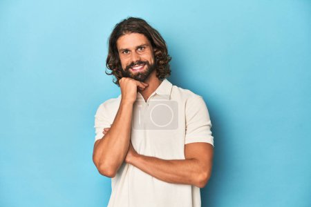Foto de Hombre de pelo largo en un polo blanco, estudio azul sonriendo feliz y confiado, tocando la barbilla con la mano. - Imagen libre de derechos