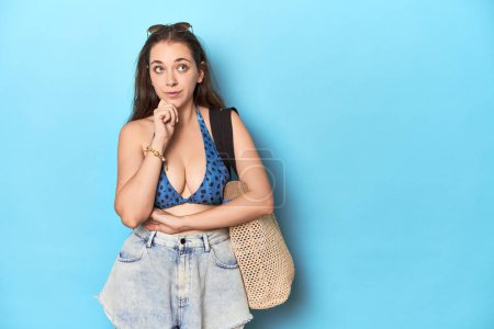 Photo for Beach-ready woman with bikini, sunglasses and bag looking sideways with doubtful and skeptical expression. - Royalty Free Image