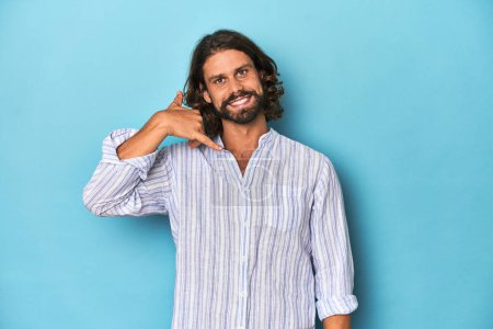 Foto de Hombre con barba en camisa de rayas azules, estudio azul mostrando un gesto de llamada de teléfono móvil con los dedos. - Imagen libre de derechos