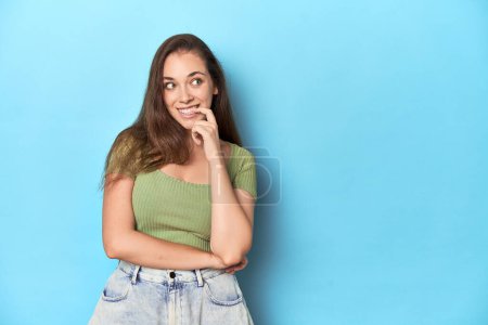 Foto de Joven mujer caucásica en un top verde sobre un fondo azul relajado pensando en algo mirando un espacio de copia. - Imagen libre de derechos