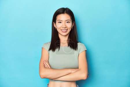 Photo for Asian woman in summer green top, studio backdrop, who feels confident, crossing arms with determination. - Royalty Free Image