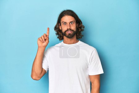 Foto de Hombre barbudo con una camisa blanca, telón de fondo azul que tiene una gran idea, concepto de creatividad. - Imagen libre de derechos