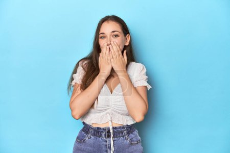 Téléchargez les photos : Jeune femme élégante en chemisier blanc sur fond de studio bleu riant de quelque chose, couvrant la bouche avec les mains. - en image libre de droit