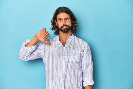 Photo for Man with beard in blue striped shirt, blue studio showing a dislike gesture, thumbs down. Disagreement concept. - Royalty Free Image