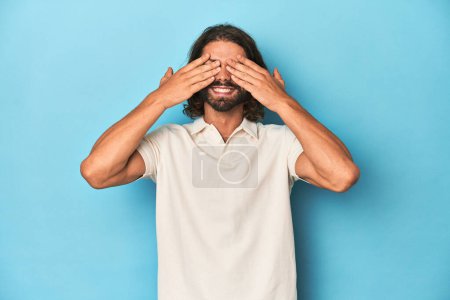 Photo for Long-haired man in a white polo, blue studio covers eyes with hands, smiles broadly waiting for a surprise. - Royalty Free Image