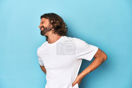 Foto de Hombre barbudo con una camisa blanca, fondo azul sufriendo un dolor de espalda. - Imagen libre de derechos