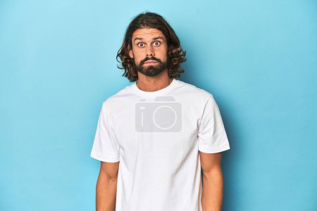Foto de Hombre barbudo con camisa blanca, fondo azul se encoge de hombros y ojos abiertos confundido. - Imagen libre de derechos