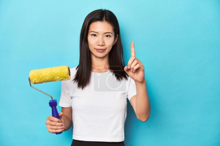 Foto de Mujer asiática joven con rodillo de pintura, concepto de bricolaje, mostrando el número uno con el dedo. - Imagen libre de derechos